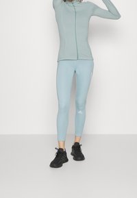 Světlomodrá sportovní bunda na zip s odpovídajícími legínami a černými teniskami, vybavená bezešvým designem a logem značky na noze.