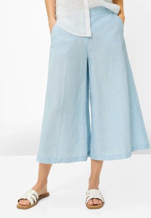 Person iført lyseblå vidde, korte bukser, hvid skjorte stoppet ind og hvide slip-on sandaler står med hænderne i lommerne.