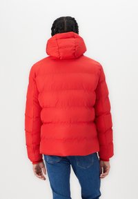 Rød puffjakke med hætte, der har et quiltet design og en elegant finish. Kombinationen med blå jeans viser en afslappet pasform og opfoldede ærmer.