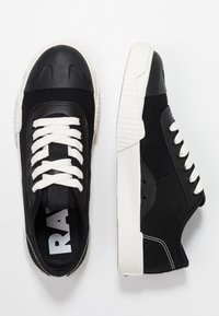 Baskets en toile noires avec lacets blancs, bout en caoutchouc, semelle texturée et coutures contrastées. Design simple et silhouette basse.