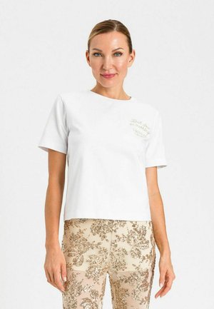 Donna con capelli pettinati all'indietro indossa una maglietta bianca semplice a maniche corte e pantaloni beige con motivi floreali di paillettes dorate su sfondo bianco.