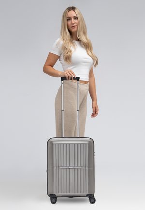 Valise grise extensible avec un design nervuré, poignée noire et quatre roues, tenue par une personne portant un haut blanc et un pantalon beige.