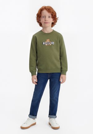 Rødtoppa barn som står, iført en olivengrønn genser med et bamseprint, blå jeans og hvite joggesko mot en hvit bakgrunn.