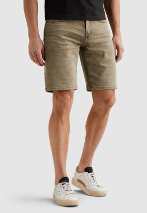 Mężczyzna w jasnobrązowych szortach dżinsowych, czarnej koszulce i białych trampkach z beżową podeszwą stojący na białym tle.