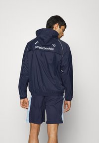 Jūras flotei raksturīgs vējjakas apvalks ar kapuci, elastīgiem piedurknēm un apakšmalu. "Sergio Tacchini" logo uz augšējās aizmugures. Saskaņots ar jūras flotes šortiem un gaiši ziliem akcentiem.