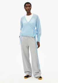 Lichtblauwe wikkeltrui van zachte stof, gecombineerd met losse grijze joggingbroek en zwarte sneakers met oranje accenten.