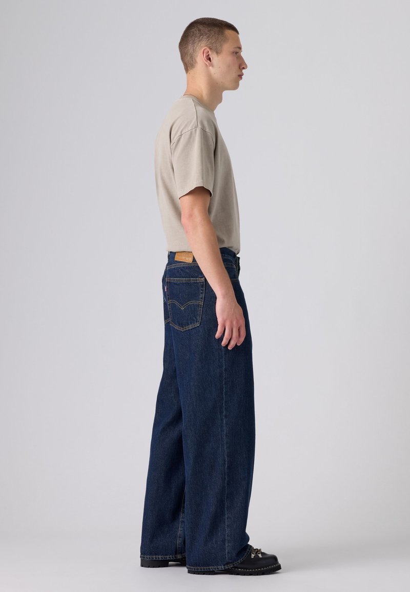 Jeune homme de profil portant un t-shirt beige, un jean large foncé et des chaussures noires sur un fond clair uni.