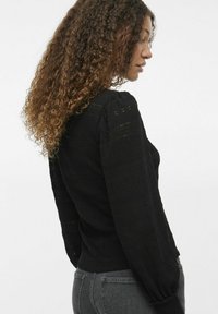 Svart stickad tröja med en åtsittande design, långa ärmar, texturerat mönster och lätt puffade axlar, i kombination med grå jeans.