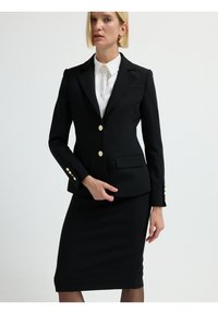 Blazer negro con botones dorados, con un corte entallado, hombros estructurados y dos bolsillos frontales. Combinado con una falda hasta la rodilla.