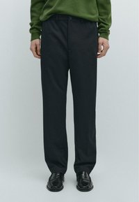 Pantaloni grigio scuro a righe sottili con taglio dritto, dotati di chiusura frontale con bottoni e passanti per cintura. Indossati con un maglione verde e scarpe nere.