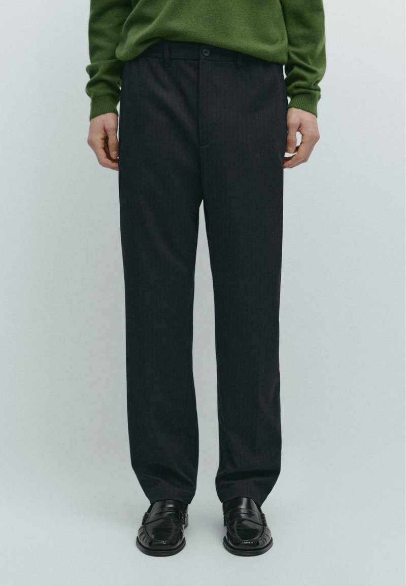 Pantaloni grigio scuro a righe sottili con taglio dritto, dotati di chiusura frontale con bottoni e passanti per cintura. Indossati con un maglione verde e scarpe nere.