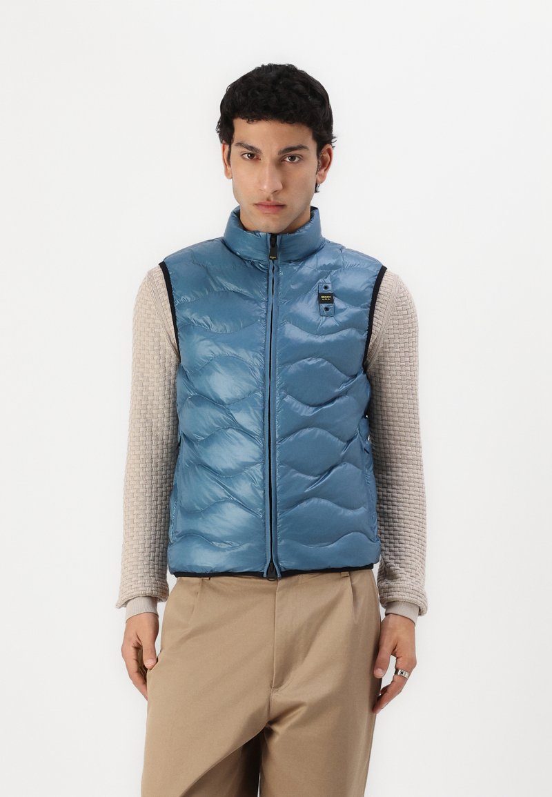 Blå puffervest med quiltet design, høy krage, frontglidelås og en liten logo detaljer. Brukt over en teksturert beige topp.
