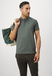 Kurzärmeliges Poloshirt mit Reißverschlusskragen aus dunkelgrünem Stoff, mit einem dezenten Logo auf der Brust. Das Modell trägt eine große grüne und cremefarbene Tragetasche.