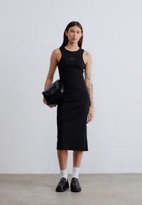 Μαύρο αμάνικο φόρεμα με εφαρμοστή γραμμή, υφή υφάσματος και διακριτική λεπτομέρεια λογότυπου· συνδυάζεται με μαύρη τσάντα και κάλτσες με παπούτσια.