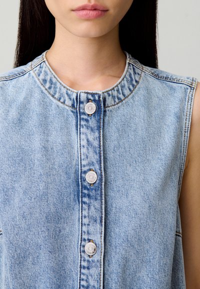 Camisa sin mangas de mezclilla azul claro con escote redondo, que presenta cinco botones a presión plateados y detalles de costura visibles.