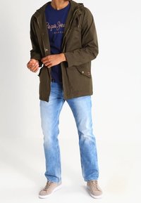 Grön lättviktsjacka med huva, parat med en marinblå t-shirt, ljusblå jeans och beige sneakers. Enkelt mönster, avslappnad passform.