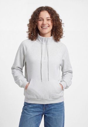 Persona sorridente con capelli ricci che indossa una felpa grigio chiaro con cerniera a un quarto e jeans blu, mani nelle tasche anteriori.