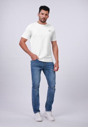 White cotton t-shirt with a small bicycle graphic on the left chest, paired with distressed light blue jeans and white sneakers.