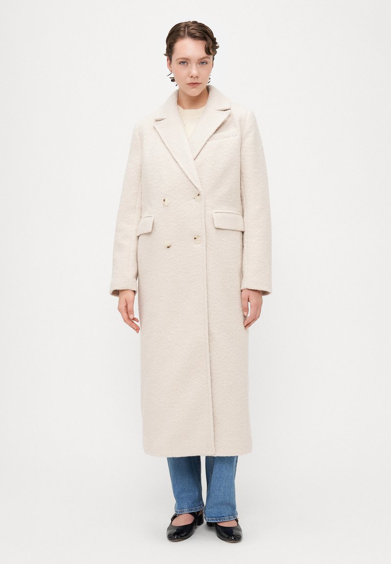 Hvit, teksturert, dobbeltspent frakk med notched lapels, sidelommer og knappdetaljer. Kombinert med blå jeans og svarte sko.