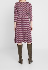 Vestido impreso con un patrón triangular en tonos burdeos y crema; cuenta con mangas tres cuartos y un dobladillo acampanado. Se lleva con botas hasta la rodilla.