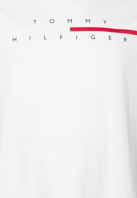 Camiseta blanca de algodón con "TOMMY HILFIGER" en texto negro y una franja de acento horizontal roja en el centro. Textura suave.