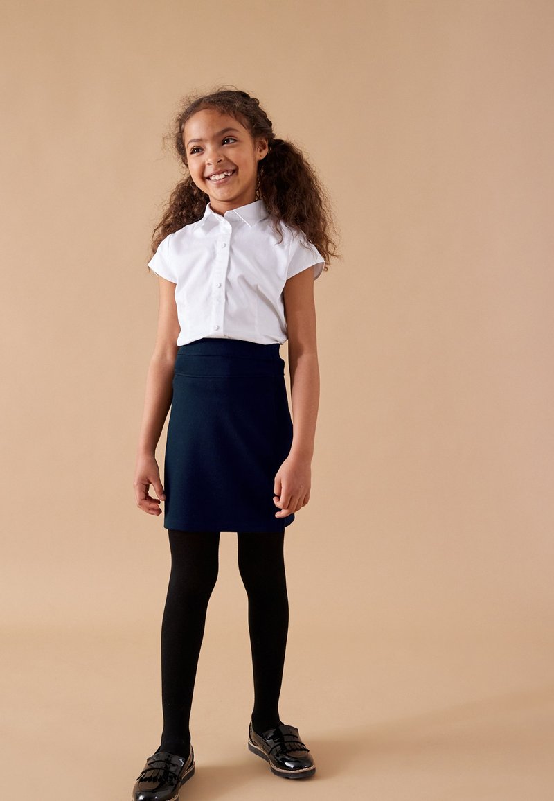Biała koszula z krótkim rękawem zapinana na guziki, granatowa spódnica trapezowa, czarne rajstopy i czarne lakierowane buty z frędzlami. Prosty design.