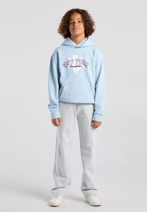 Kind met krullend haar dat een lichtblauwe New York hoodie draagt, grijze joggingbroek en witte sneakers, staand tegen een effen achtergrond.