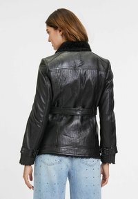 Zwarte leren jas met een nepkraag van bont, een ceintuur in de taille en een gestructureerde afwerking. Voorzien van zilveren hardware op de mouwen en een gladde achterkant.