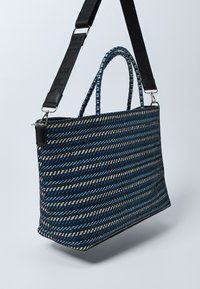 Obdélníková tkaná taška přes rameno s modrými, černými a béžovými diagonálními pruhy, vybavená pletenými držadly a černým nastavitelným ramenním popruhem.