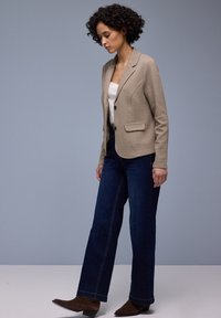 Vrouw met krullend haar, gekleed in een beige gebreide blazer, een wit topje, donkerblauwe wijde jeans en bruine enkellaarsjes, staand tegen een effen achtergrond.