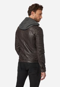 B brown läderjacka med quiltade detaljer på axlarna, med en grå hoodie fäst. Slät textur, figursydd design och tryckknappsstängning vid ärmsluten.