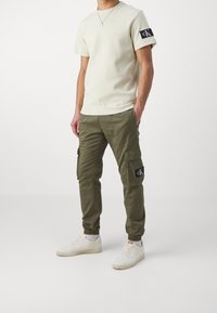 Beige short-sleeve t-shirt with a textured fabric and black logo patch, paired with olive cargo pants featuring side pockets and elastic cuffs.