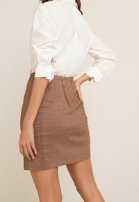 Femme portant un chemisier blanc à manches longues retroussées et une mini-jupe brune à carreaux taille haute avec une fermeture éclair visible au dos.