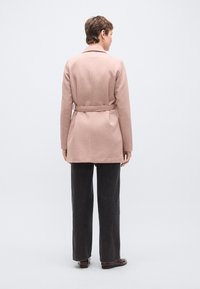 Φωτεινό ροζ παλτό με ζώνη και δομημένο σχέδιο, μακριά μανίκια και γιακά, σε συνδυασμό με σκούρο παντελόνι φαρδιάς γραμμής, απεικονισμένο από πίσω.