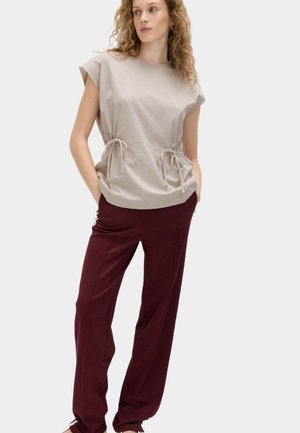 Donna con capelli ricci che indossa un top beige senza maniche con lacci laterali e pantaloni larghi color borgogna, in piedi con le mani nelle tasche su sfondo bianco.