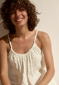 Débardeur blanc texturé avec des bretelles fines, présentant un tissu froncé et un col lisse. Le mannequin sourit devant un fond neutre.