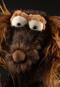 Stuffed animal resembling a dog, with dark brown fur, light brown accents around the eyes, and a round, brown, textured nose.