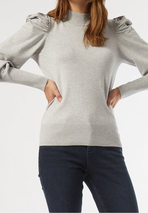Femme portant un pull gris clair à manches bouffantes et un jean bleu foncé, les mains posées sur les hanches, sur fond blanc.