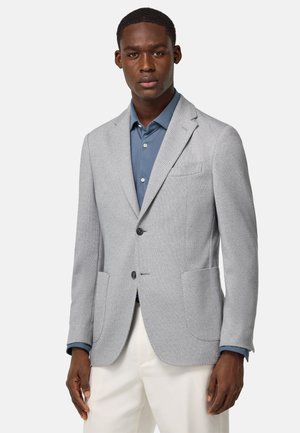 Hellgrauer Blazer aus strukturiertem Stoff. Mit Reverskragen, zwei Vorderentaschen und zwei Knöpfen. Über einem blauen Hemd getragen.