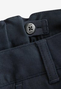 Mörkblå byxor med en robust midja, en silverknapp med fyra hål, och en subtilt texturerad tyg. Clarks-logotyp synlig på knappen.