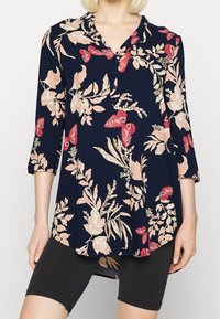 Tunic blu navy con stampa floreale rosa e farfalle rosse, maniche a tre quarti, scollatura a V con bottoni, indossata con pantaloni corti neri aderenti.