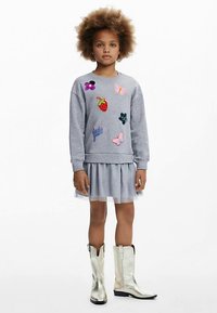 Graues Sweatshirt mit bunten, bestickten Abzeichen, kombiniert mit einem hellgrauen Tüllkleid und silbernen Cowboy-Stiefeln.