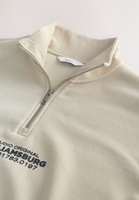 Hellbeiger Pullover mit Reißverschlusskragen, geripptem Kragen und dunkelblauem Text "STUDIO ORIGINAL WILLIAMSBURG 01783.0197." Weiche Textur.