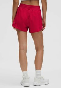 Raudoni sportiniai šortai su virvele juosmens srityje, pasižymintys glotnia tekstūra ir šiek tiek lenktu kraštu. Dėvimi su baltais sportiniais batais ir kojinėmis.