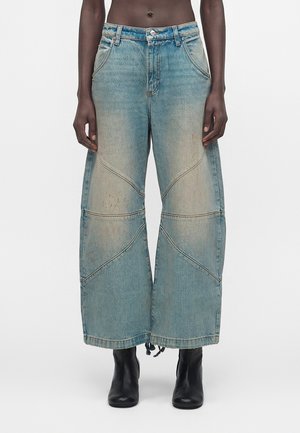 Blå jeans med vide ben, falmede paneler og buede sømdetaljer, båret med sorte ankelstøvler på en person, der står mod en ensfarvet baggrund.