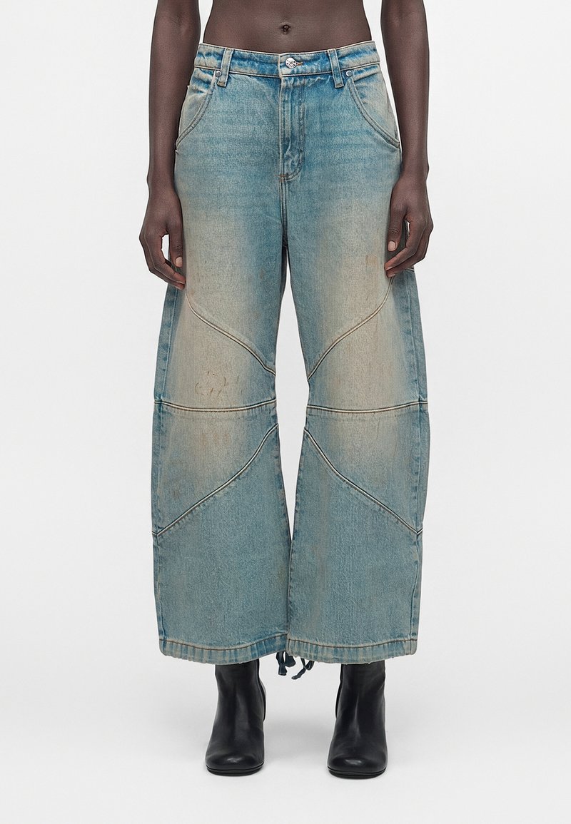 Μπλε τζιν παντελόνι με φαρδιά μπατζάκια, ξεθωριασμένες λεπτομέρειες και καμπυλωτές ραφές, φορεμένο με μαύρα μποτάκια αστραγάλου σε άτομο που στέκεται μπροστά σε απλό φόντο.