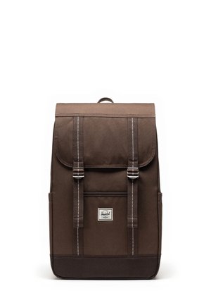 Mochila de lona marrón con una solapa, dos correas frontales y forma rectangular. Presenta un pequeño parche de logo en la parte delantera y bolsillos laterales.