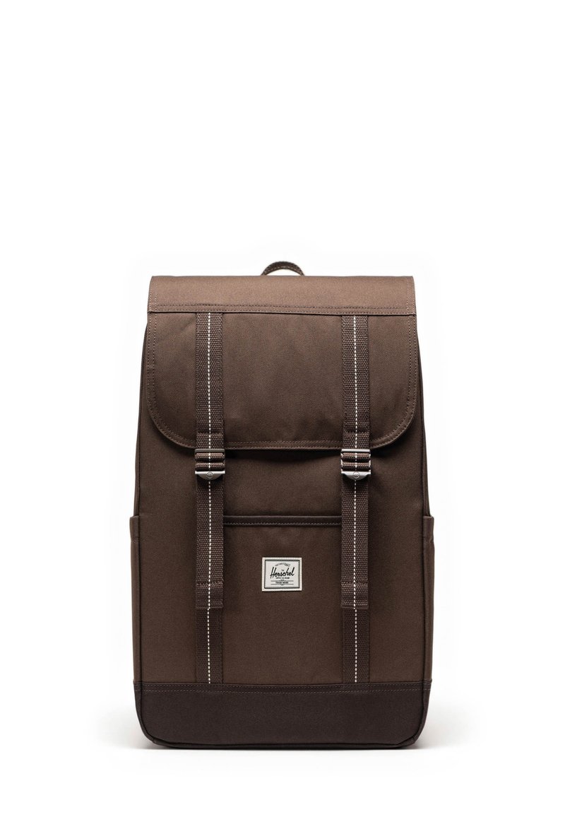 Sac à dos en toile marron avec un rabat, deux sangles à l'avant et une forme rectangulaire. Il présente un petit patch logo à l'avant et des poches latérales.
