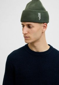 Groene geribbelde gebreide beanie met gestikte label, gedragen met een marineblauwe gestructureerde trui. Focus op de vorm en materiaaldetails van de muts.