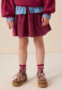 Enfant portant une jupe en velours côtelé bordeaux, un sweat-shirt rouge à motifs de cœurs, des chaussettes en léopard roses, et des baskets marron avec des lacets roses.